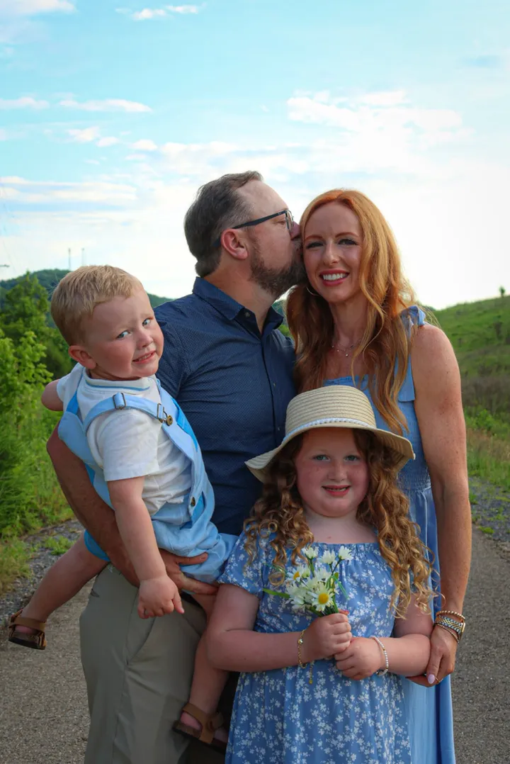 Ryan kissing Brittany on the cheek while holding Kamden, with Kinsley in front holding daisies