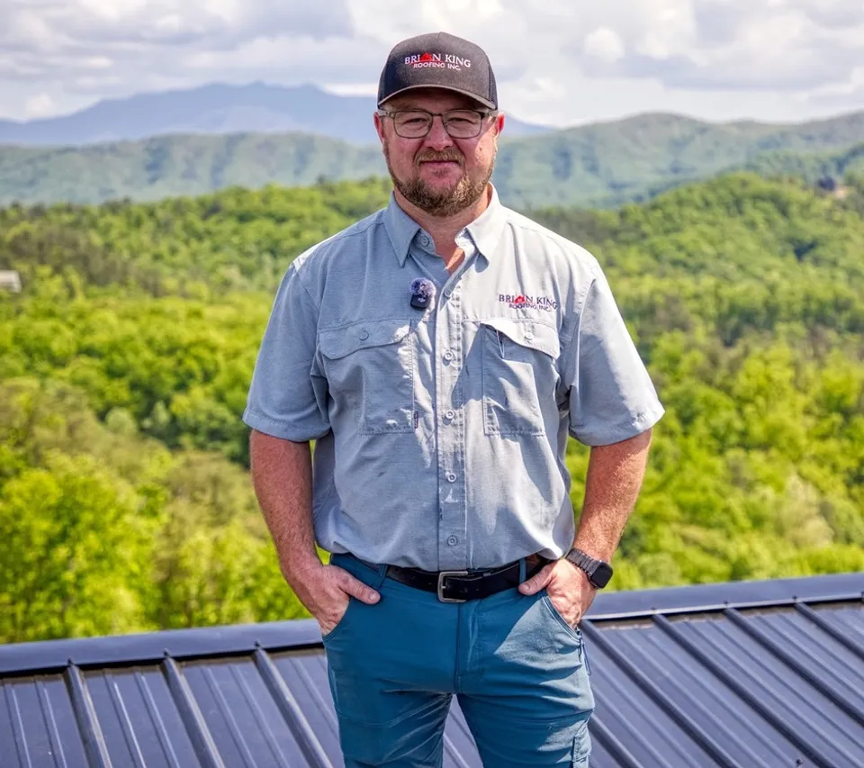 Ryan King in a Brian King Roofing shirt and cap, standing on a metal roof with the Smoky Mountains behind him