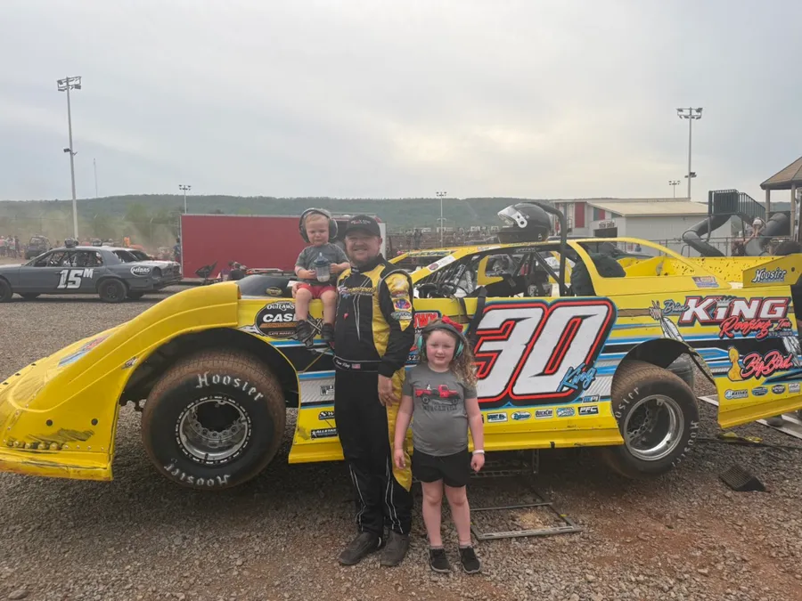 Ryan in his driver suit with Kinsley and Kamden beside the yellow #30 Brian King Roofing late model